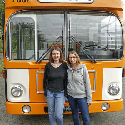 Bus, vordere Ansicht, zwei Koordinatorinnen vor dem Bus